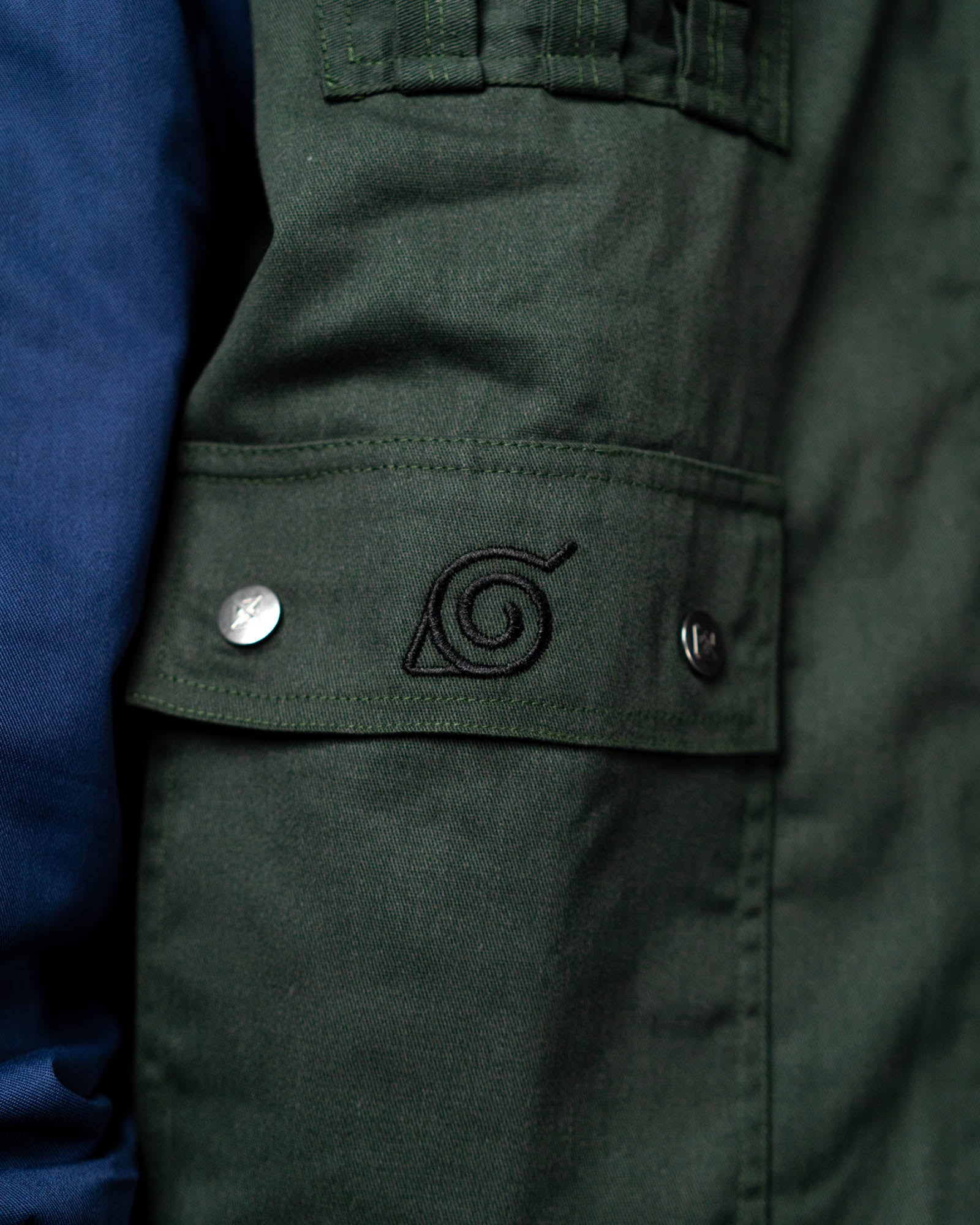 Close-up of the Naruto Shippuden Jonin Jacket pocket flap with black Hidden Leaf embroidery and silver snap buttons